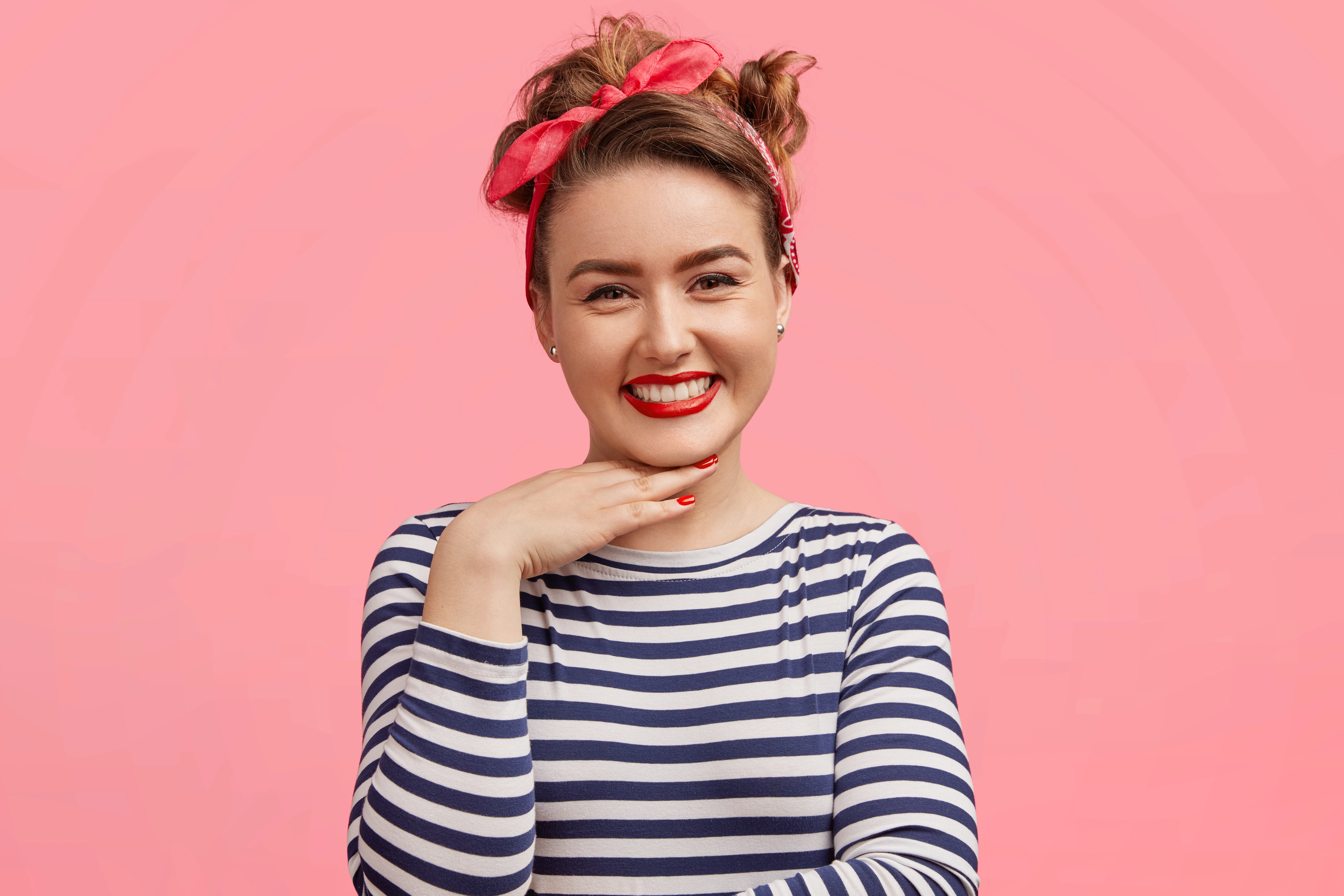 candid shot of pleasant looking female with make up has positive expression keeps hand under chin wears striped sweater and stylish headband