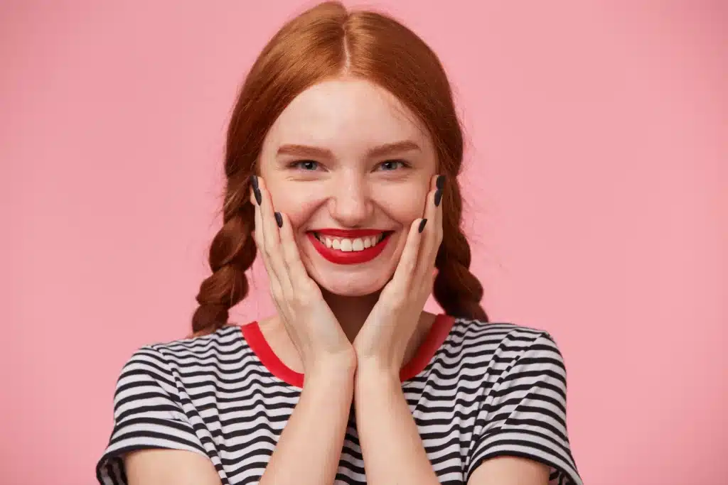 close up of glad joyful beautiful red haired girl with two braids keeps hands near her face and smiling rosily happily with red lips