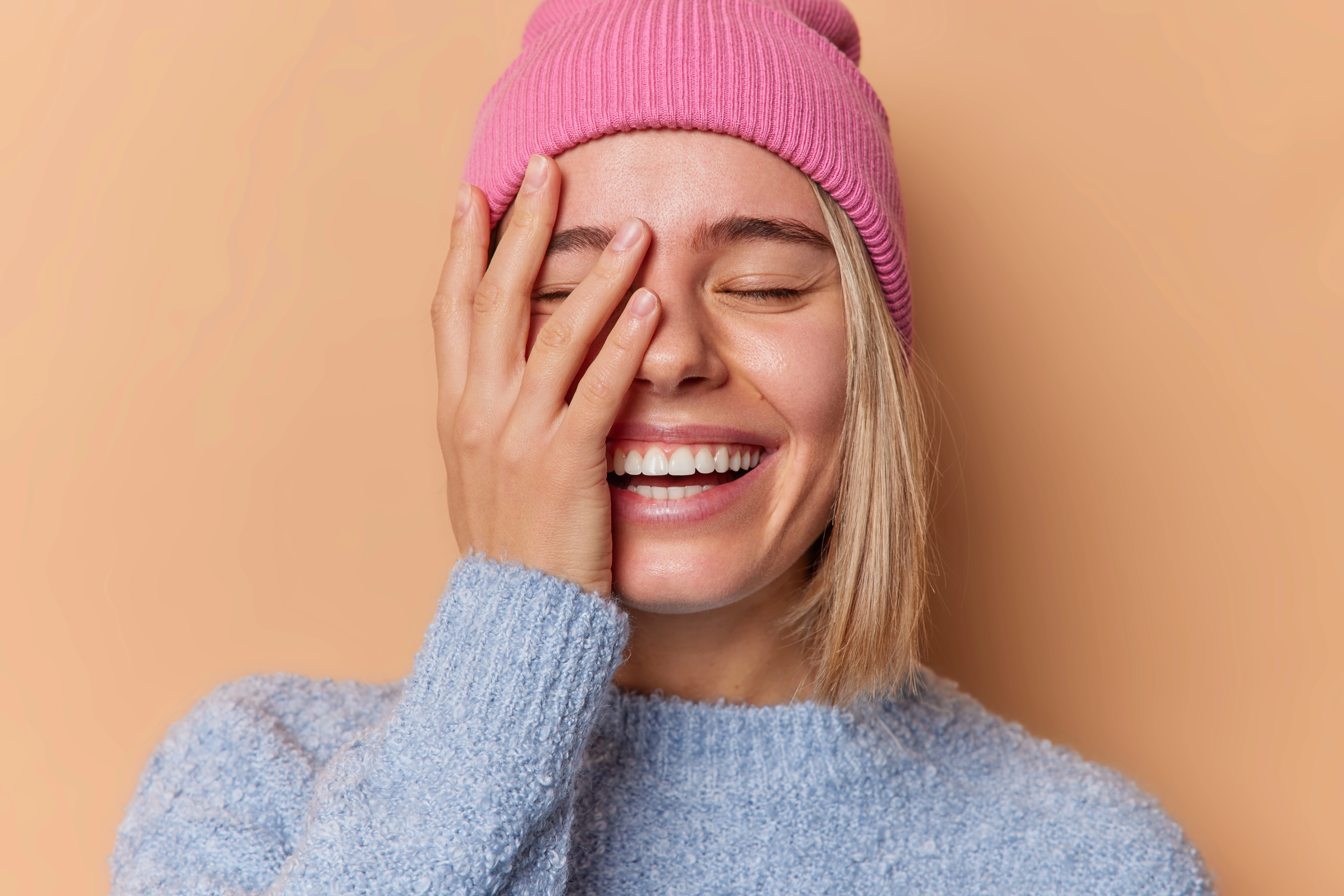 happy optimistic young woman makes face palm smiles broadly keeps eyes closed being in good mood wears hat and sweater isolated over beige background positive emotions and feelings concept