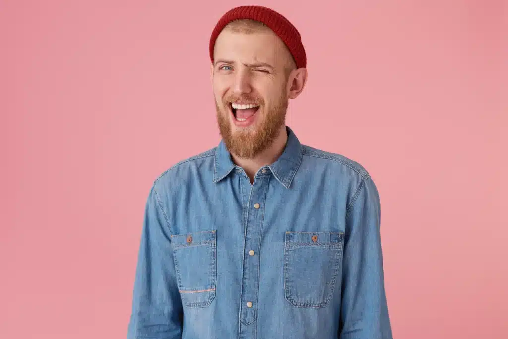 positive bearded male with fashioned denim shirt red hat has playful expression winks mouth opened encourages supports isolated human facial expressions body language
